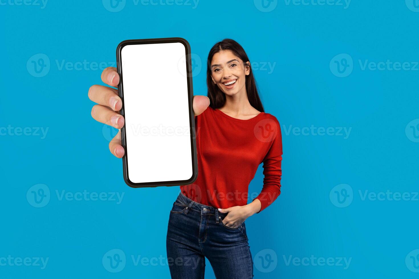 A woman wearing a red shirt is holding up a cell phone with blank screen in her hand, the screen is visible. She appears to be engaging with the device, possibly taking a photo or sending a message.