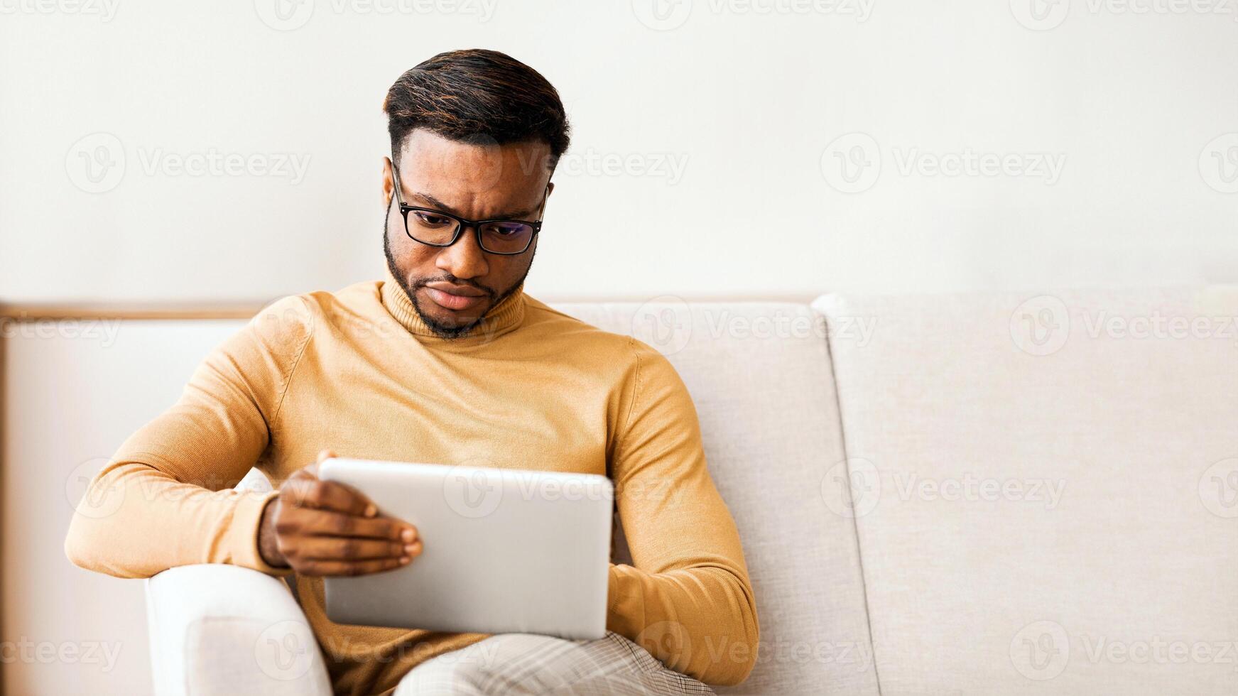 Serious Afro Entrepreneur Man Using Digital Tablet Sitting On Sofa At Home. Selective Focus photo
