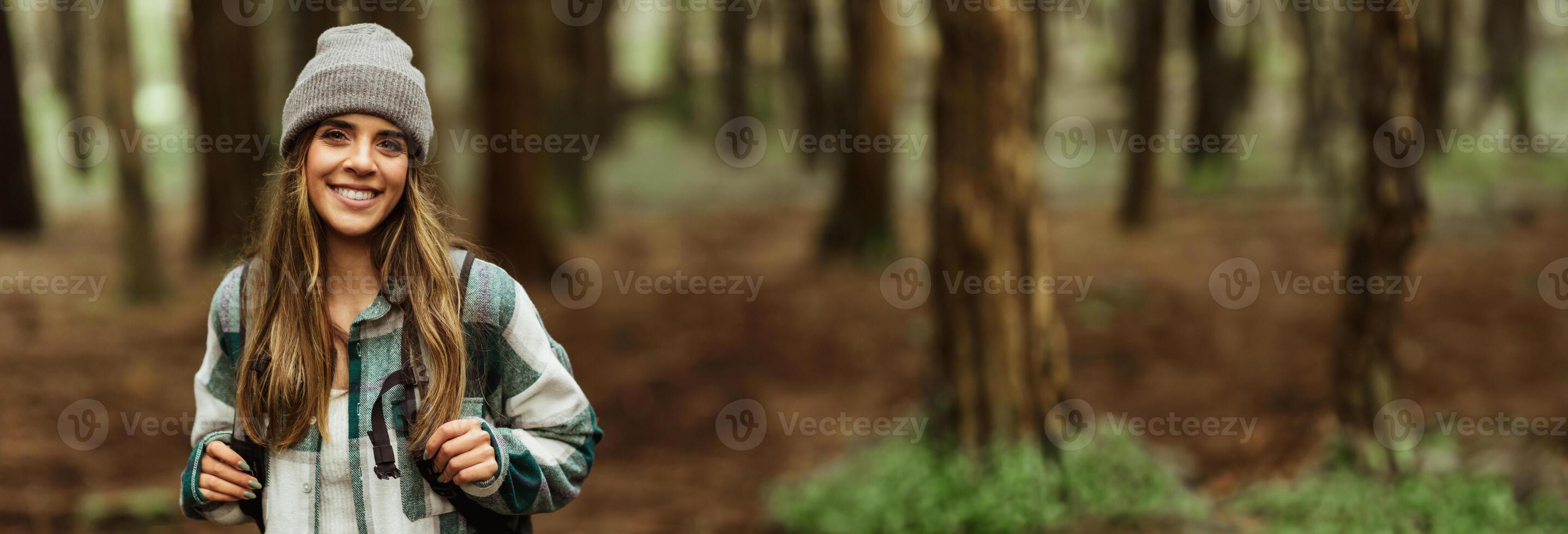 A young woman with long brown hair, wearing a grey beanie and a plaid shirt, smiles brightly as she hikes through a dense forest, copy space photo