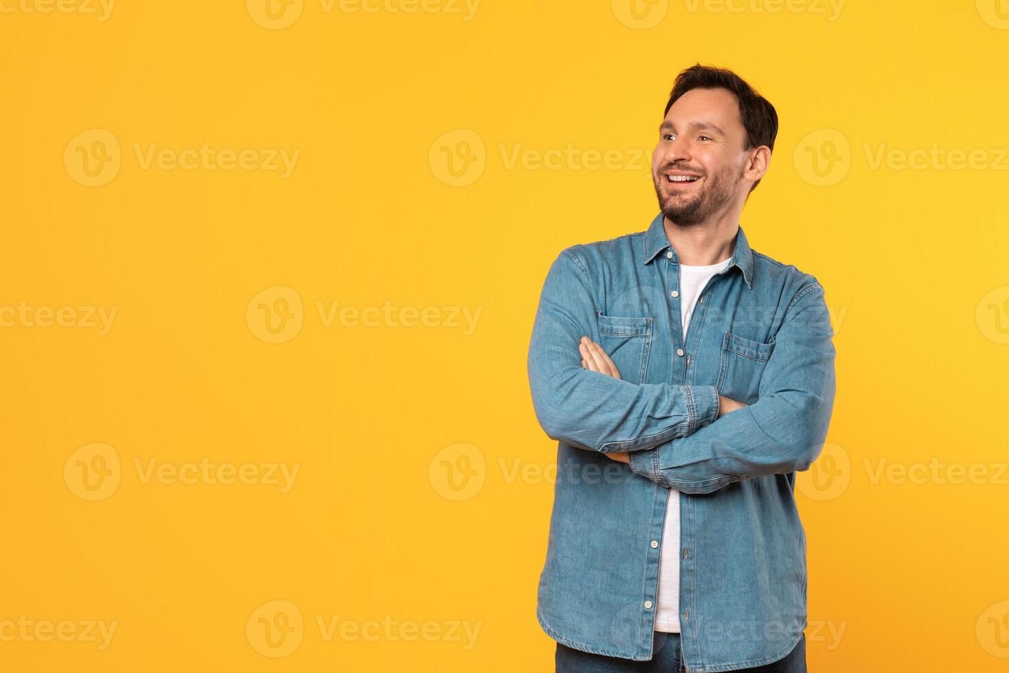 A man with a beard and short brown hair wears a blue denim shirt with a white undershirt. He stands in front of a yellow background, smiling, with his arms crossed. He is looking at copy space photo