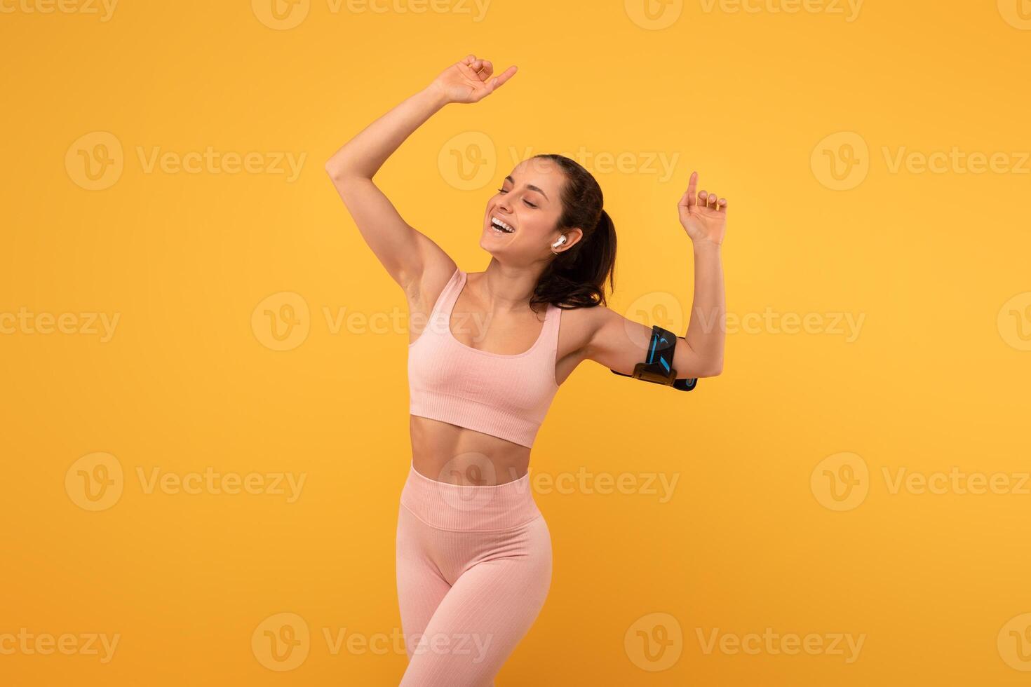 A young woman with a beaming smile is captured mid-dance against a bright yellow backdrop, dressed in form-fitting athletic wear, and is clearly reveling in a moment of triumph or celebration photo