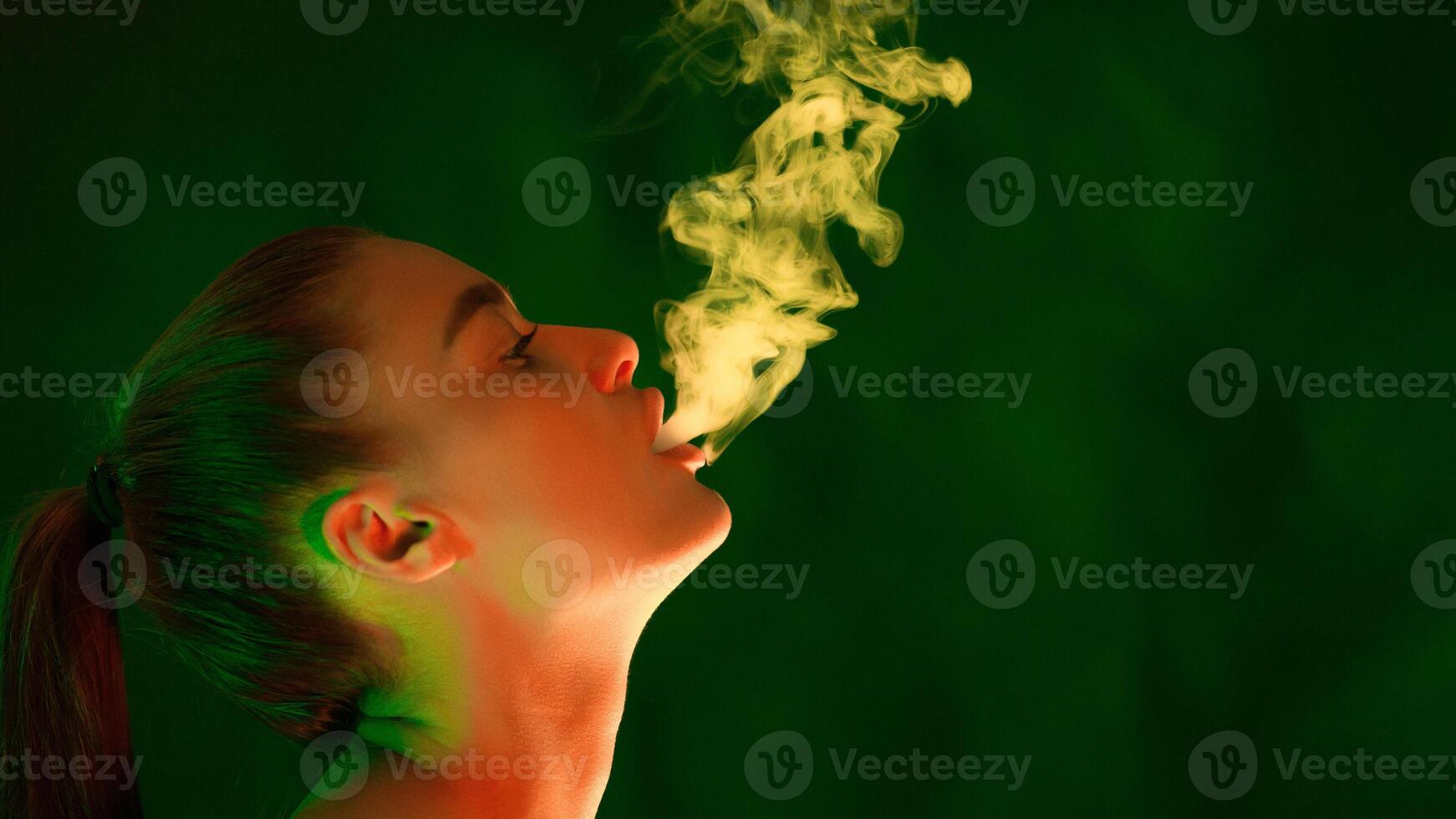 A woman is standing in front of a plain green background, smoking a cigarette. She is blowing smoke into the air, copy space photo