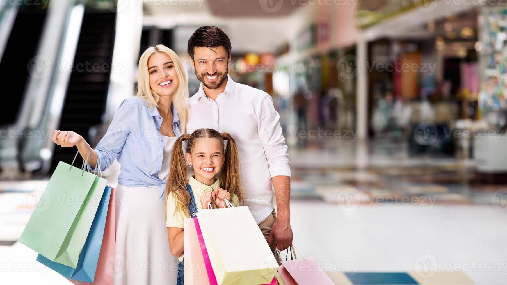 A young family, a mother, father, and daughter, are standing in a large mall. They are all smiling happily and holding shopping bags, copy space photo