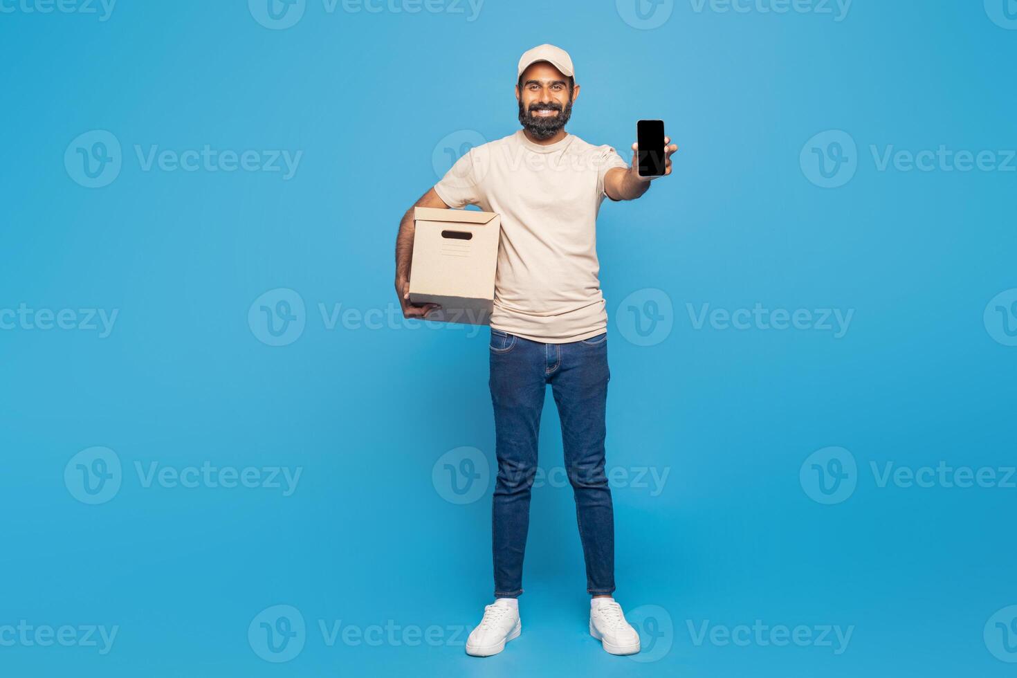 Indian delivery man is standing and smiling while holding a cardboard box in one hand and displaying a smartphone in the other photo