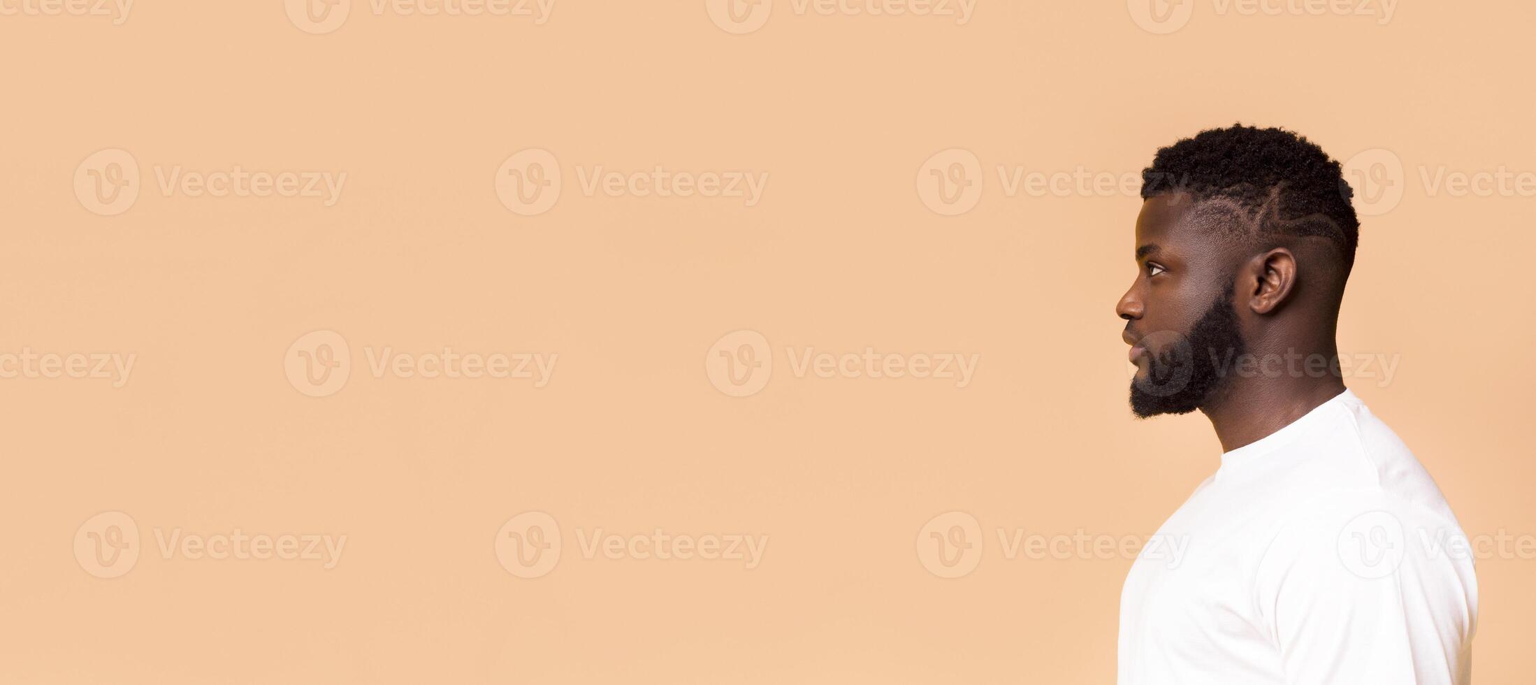 African American man with short, styled hair and a beard. He is wearing a white t-shirt and standing in front of a light peach background, panorama with copy space photo
