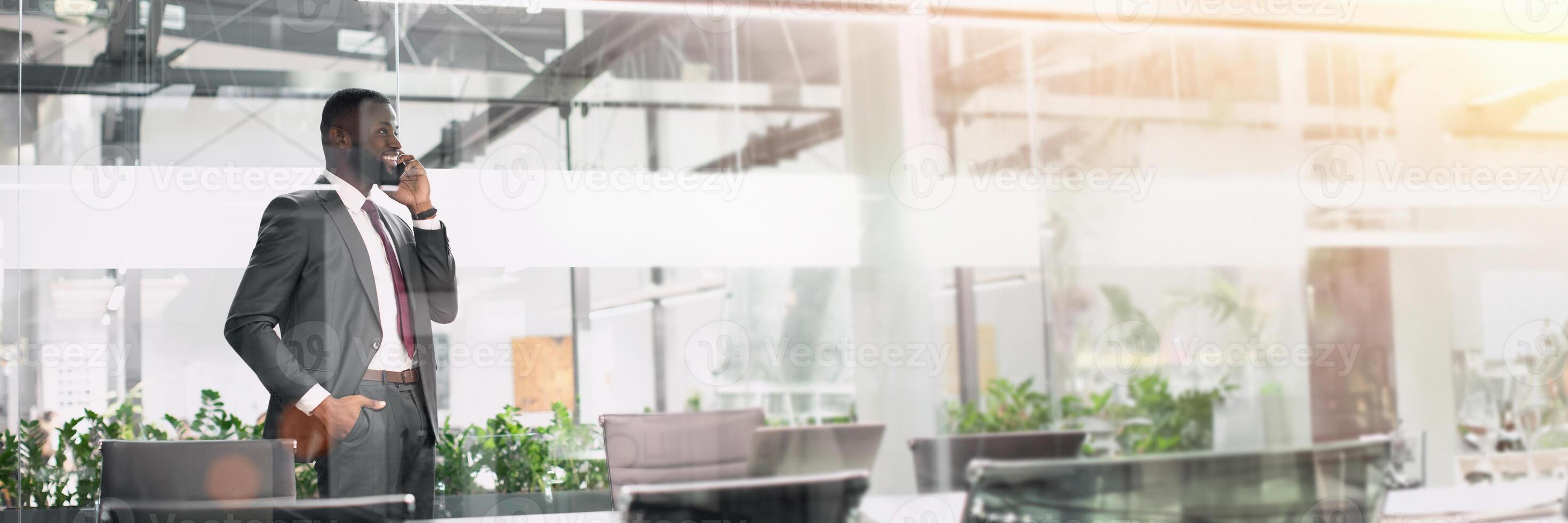 Cheerful handsome african american young businessman in nice suit talking on phone with business partner, standing in conference hall, looking at copy space and smiling, panorama photo