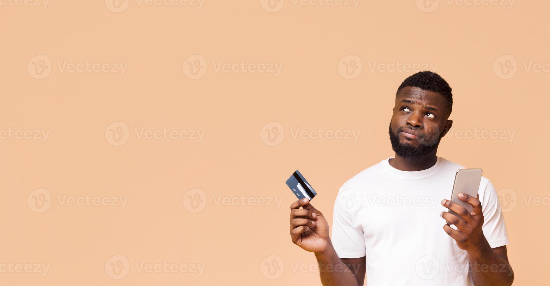 African American man in a white t-shirt stands against a light beige background, holding a credit card in his left hand and a smartphone in his right. Panorama with copy space photo