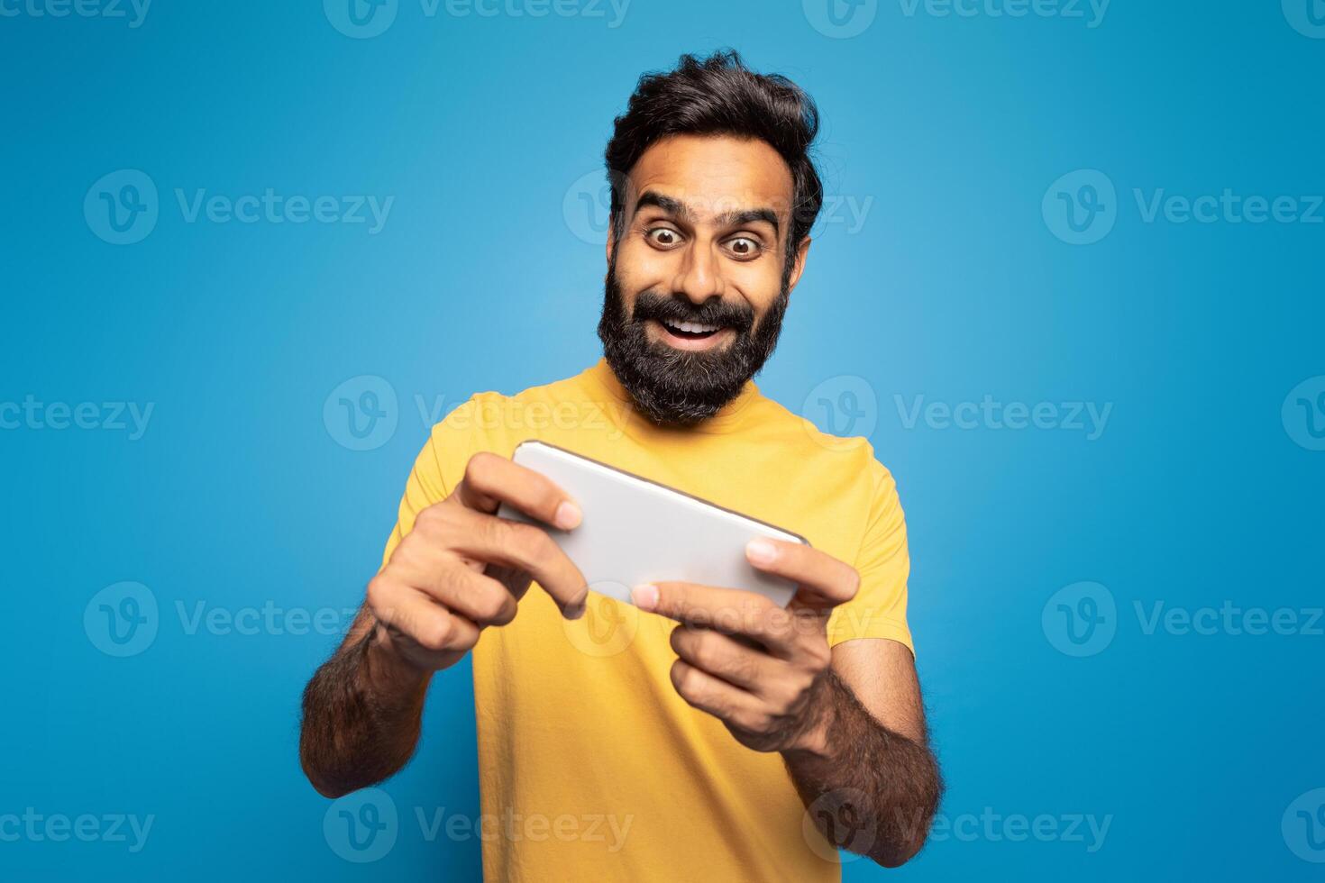 A joyful Indian man wearing a yellow shirt is enthusiastically playing a mobile game. He is set against a vibrant blue background, fully immersed in the game. photo