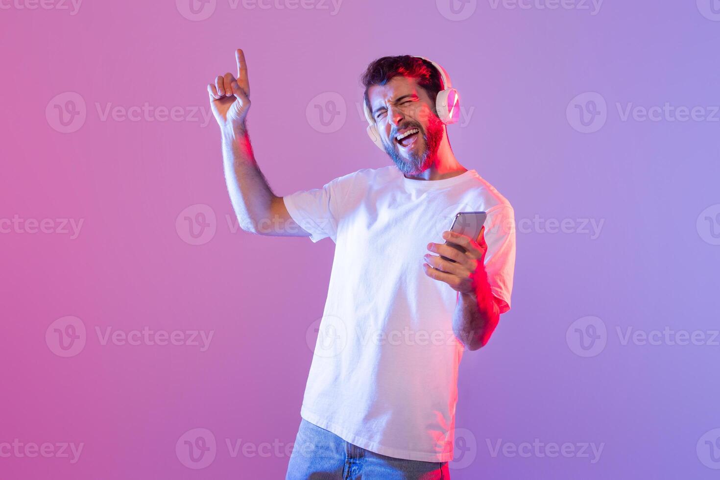 A man wearing headphones and a white t-shirt is joyfully dancing and singing while holding a smartphone. The brightly lit indoor setting features a vibrant purple and pink background photo