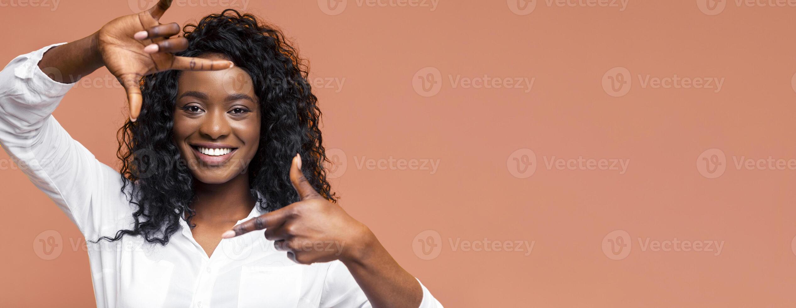 The image shows African American woman with curly black hair smiling while making a frame shape with her hands against a peach-colored background. She is wearing a white shirt. photo