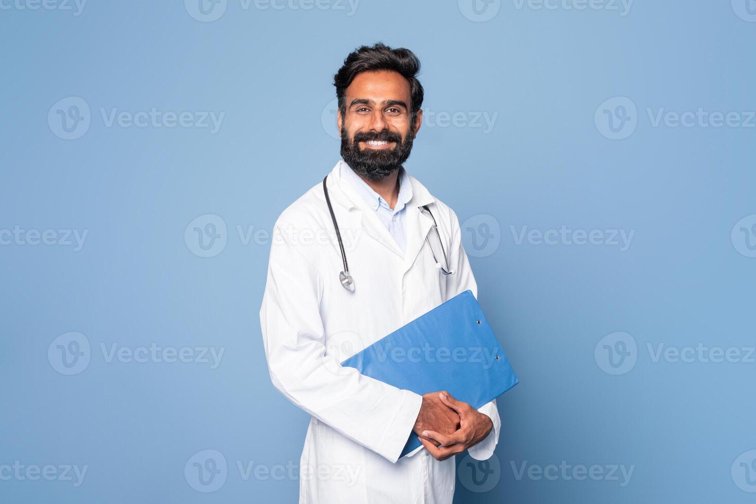 A cheerful Indian doctor is seen holding a blue folder while standing against a plain blue background. He is wearing a white lab coat and a stethoscope around his neck photo