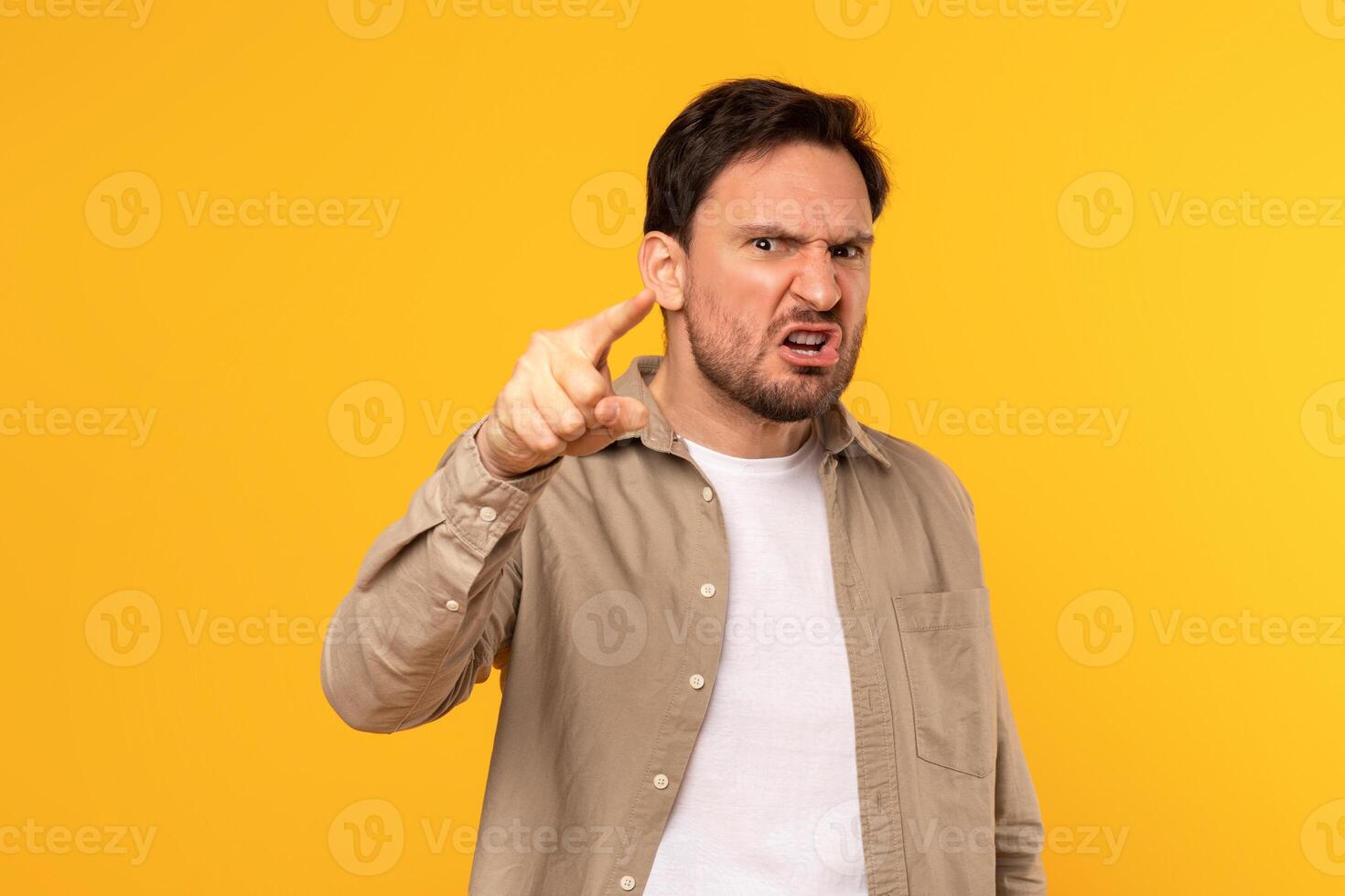A man is shown in a studio, wearing a light brown button-down shirt over a white t-shirt, looking directly at the camera with a furious expression and his index finger pointed out photo