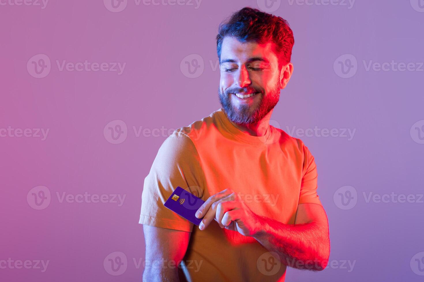 A man is standing holding a credit card in his right hand. He appears happy and relaxed, studio background with neon light photo