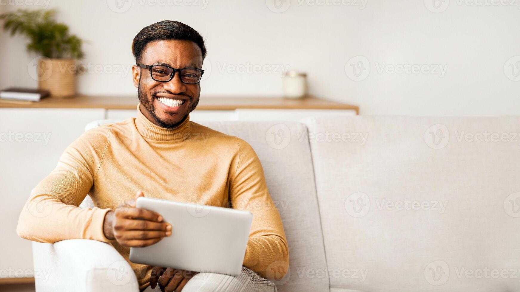 Weekend. African American Man In Eyeglasses Holding Digital Tablet And Smiling To Camera Sitting On Couch At Home. Selective Focus photo