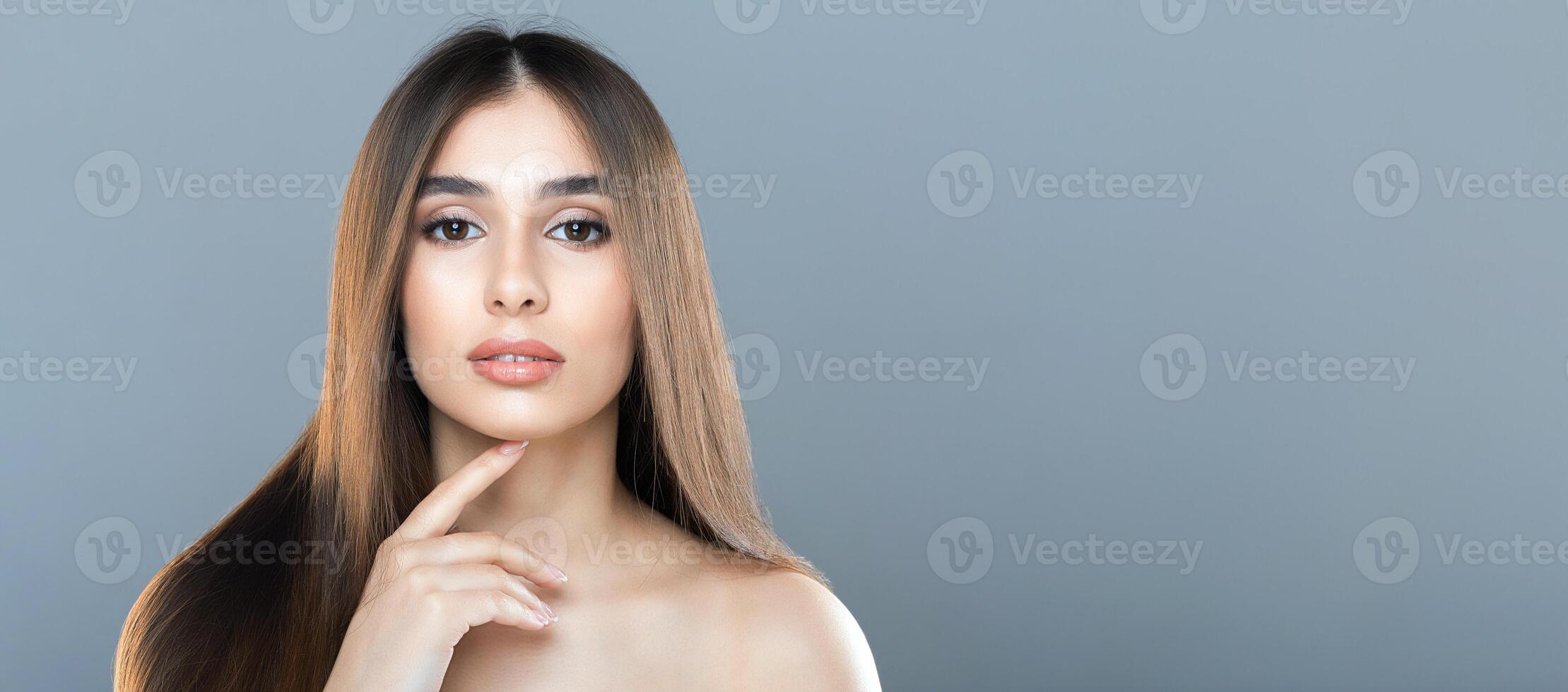 A woman with long, straight hair gazes directly at the camera with a calm expression. Her hand gently rests near her chin, and she stands against a plain gray background, copy space photo