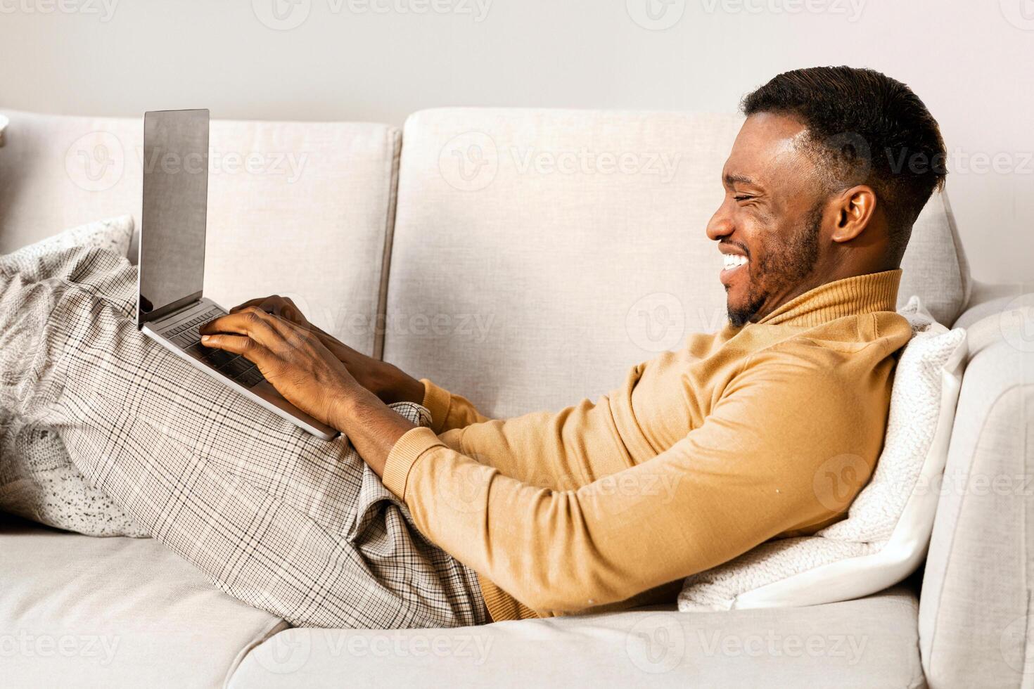 Happy African American Man Using Laptop Computer Lying On Couch Indoor. Weekend At Home Concept. photo