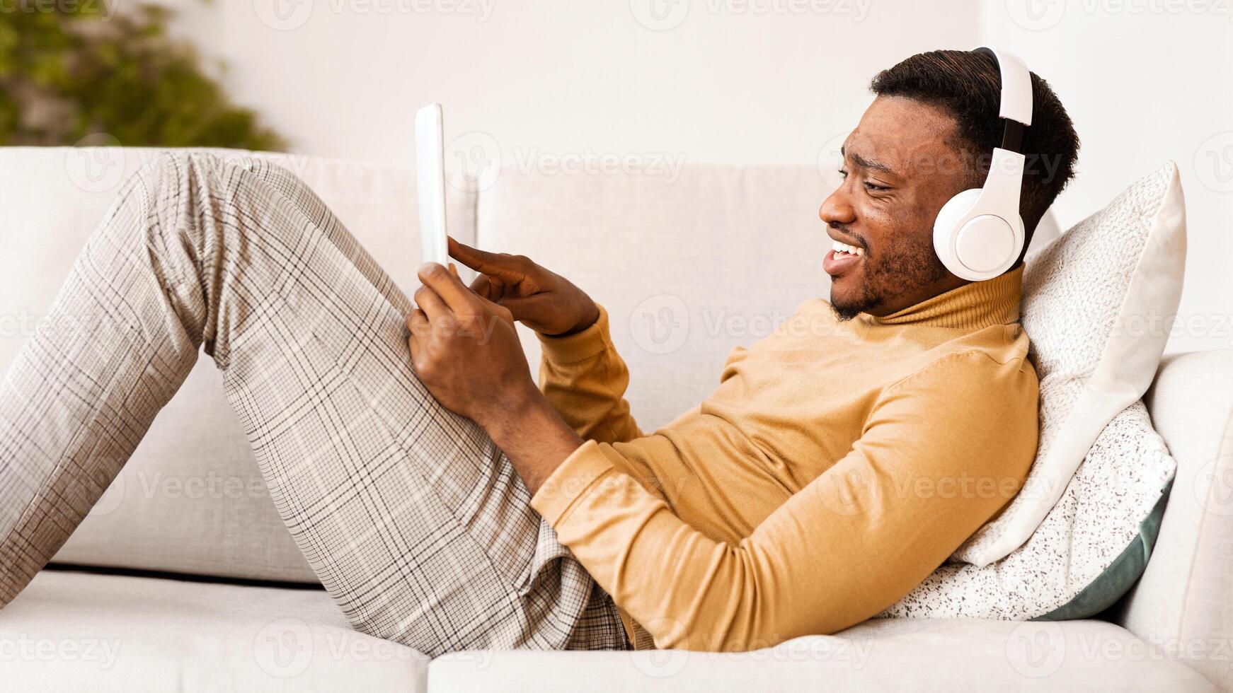 Weekend Concept. Happy African American Guy In Headphones Using Tablet Computer Listening To Music Relaxing Lying On Couch Indoor photo