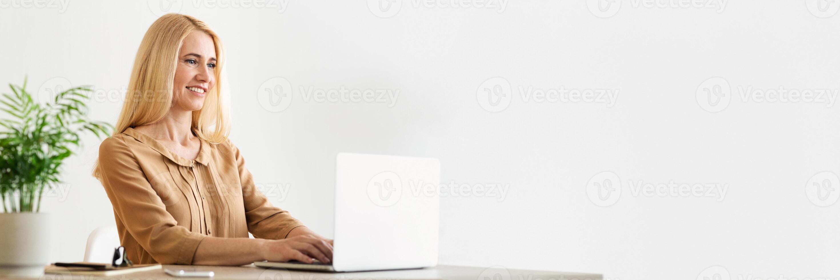 Blonde millennial business lady sitting at workplace, working on laptop at cozy office, chatting with clients, sending emails, panorama with copy space photo