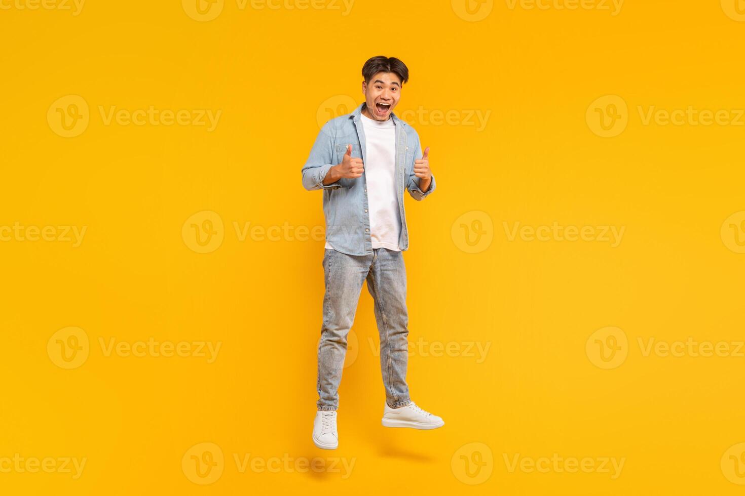 This image shows a young man in a blue shirt and jeans jumping in front of a yellow background. He is smiling and giving a thumbs up with both hands. He seems to be happy and excited about something. photo
