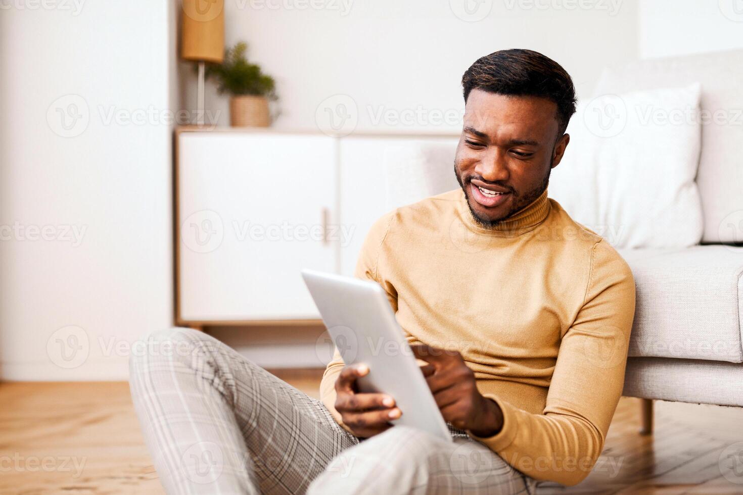 Afro Man Using Digital Tablet Reading Book Online Sitting On Floor At Home. Selective Focus photo