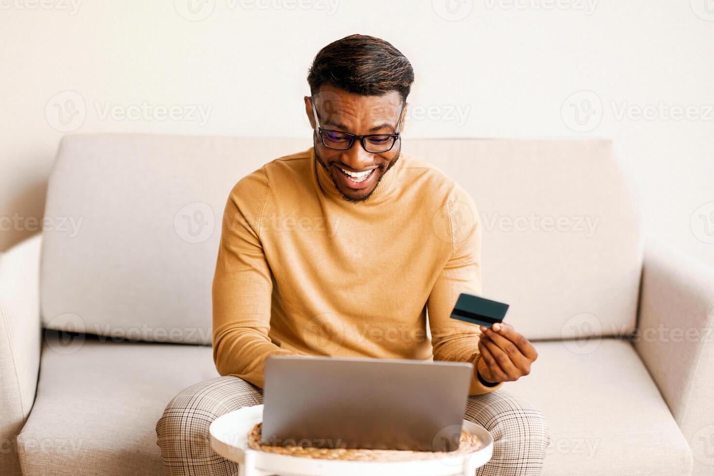 Online Shopping. Excited Black Guy Using Credit Card And Laptop Computer Sitting On Sofa Indoor. Selective Focus photo