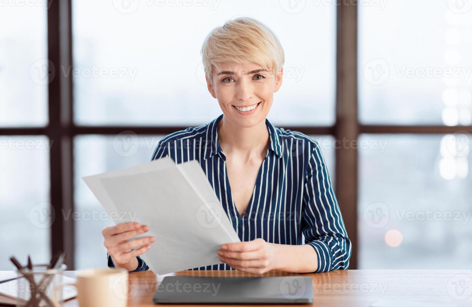 Cheerful Businesswoman Holding Documents Sitting At Laptop Smiling To Camera Working In Modern Office. Selective Focus photo
