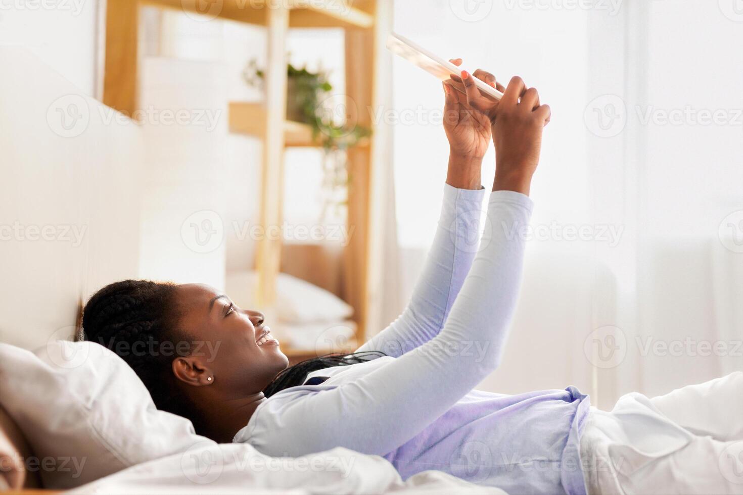 Happy African American Lady Using Tablet Computer Reading Interesting Blog Lying On Back In Bed At Home. Selective Focus photo