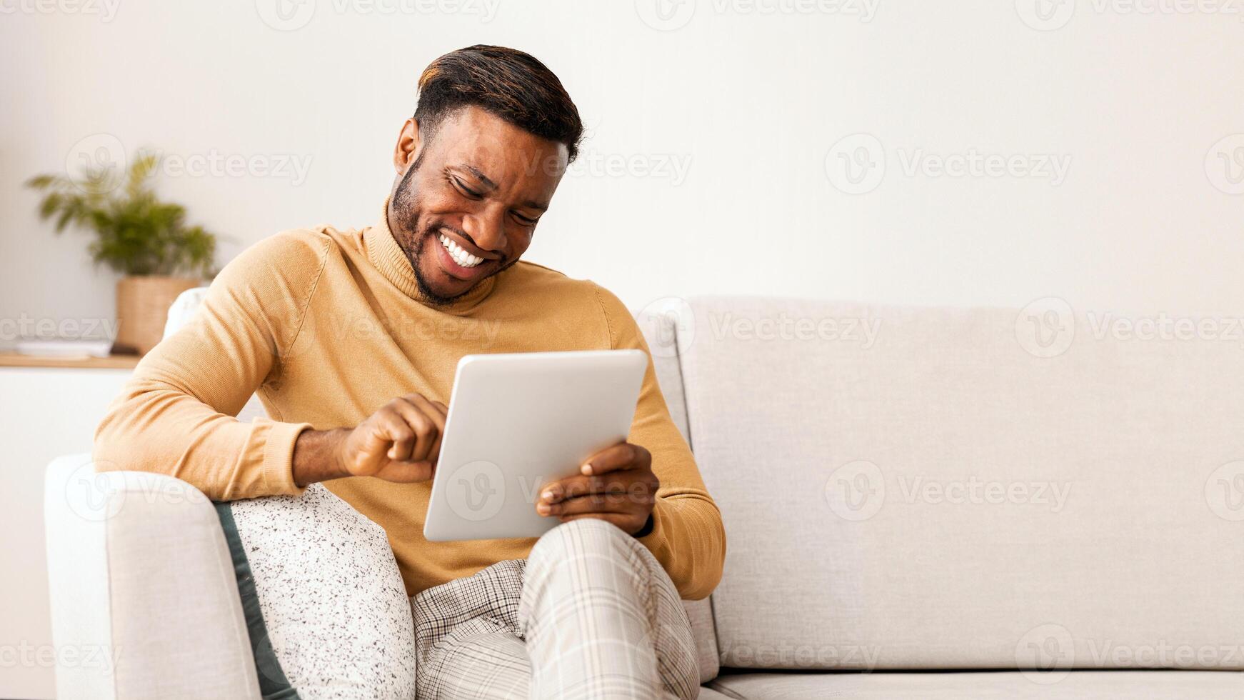 Cheerful Black Guy Using Digital Tablet Smiling Sitting On Sofa At Home. Cool Mobile Application. Selective Focus photo
