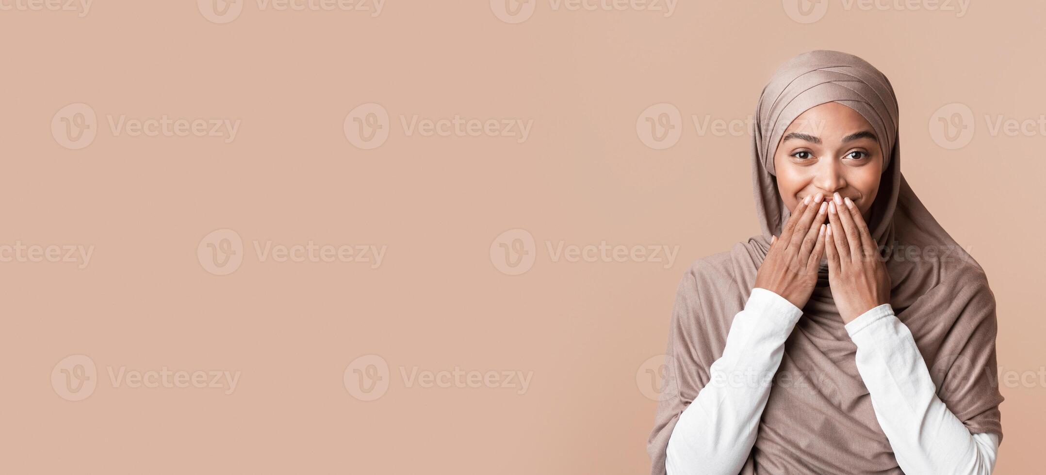 Good news. Surprised afro muslim woman in hijab covering her mouth with hands over yellow background with copy space photo