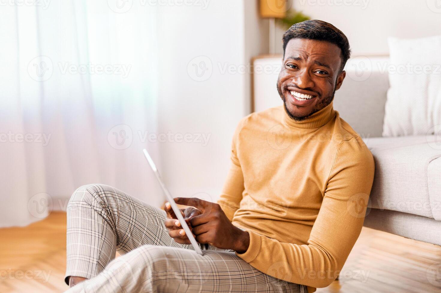 Positive Afro Man Using Tablet Computer Smiling To Camera Sitting On Floor At Home. Selective Focus photo