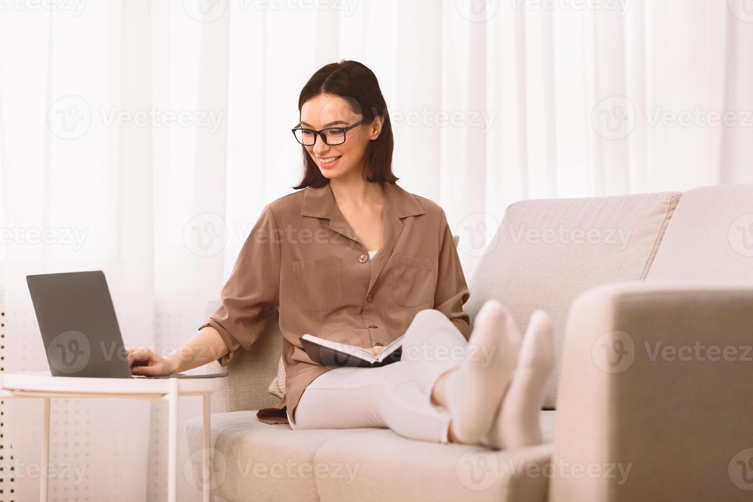 conocimiento concepto. relajado niña en lentes sentado en sofá, leyendo libro, participación cuaderno y utilizando computadora portátil, copyspace foto