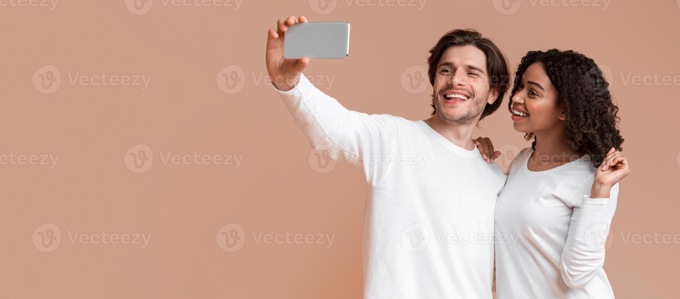 Say Cheese. Cheerful interracial couple taking sellfie on smartphone together, posing over yellow background with free space photo