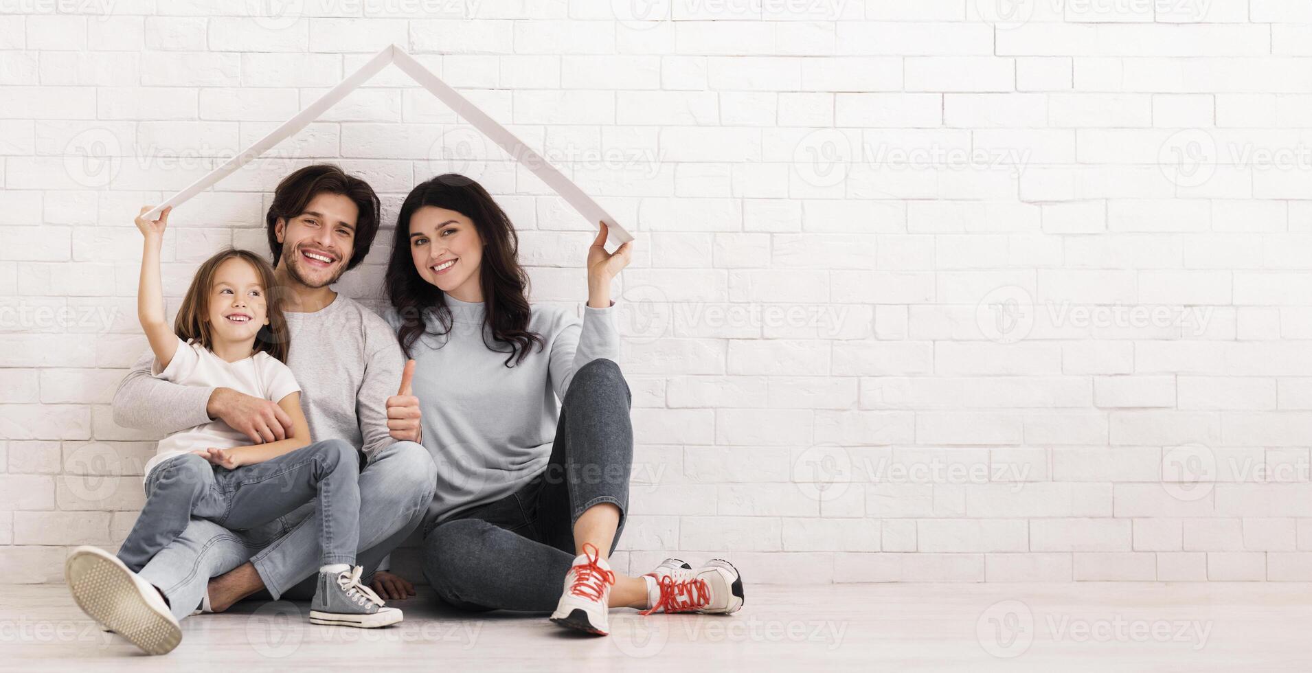 Insurance For Family Concept. Happy Parents And Little Daughter Sitting On Floor Under Symbolic Cardboard Roof And Smiling, Empty Space photo