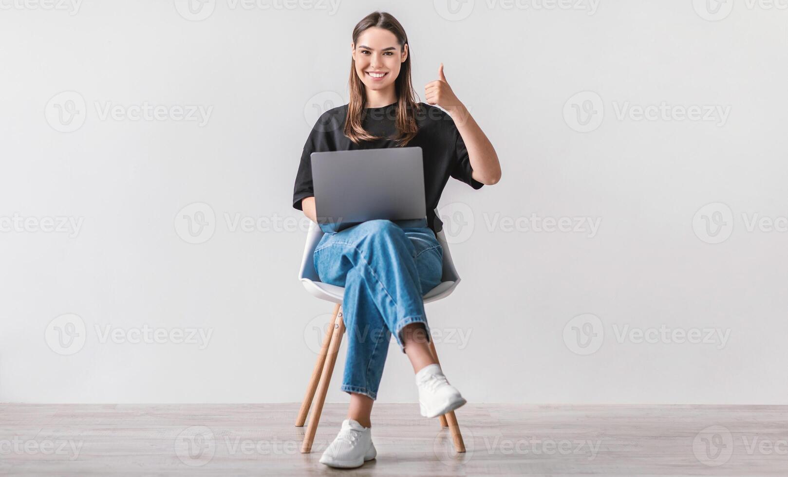 Smiling millennial woman working on laptop, showing thumb up gesture, recommending new website, sitting on chair against white wall. Young lady surfing internet, searching for information online photo