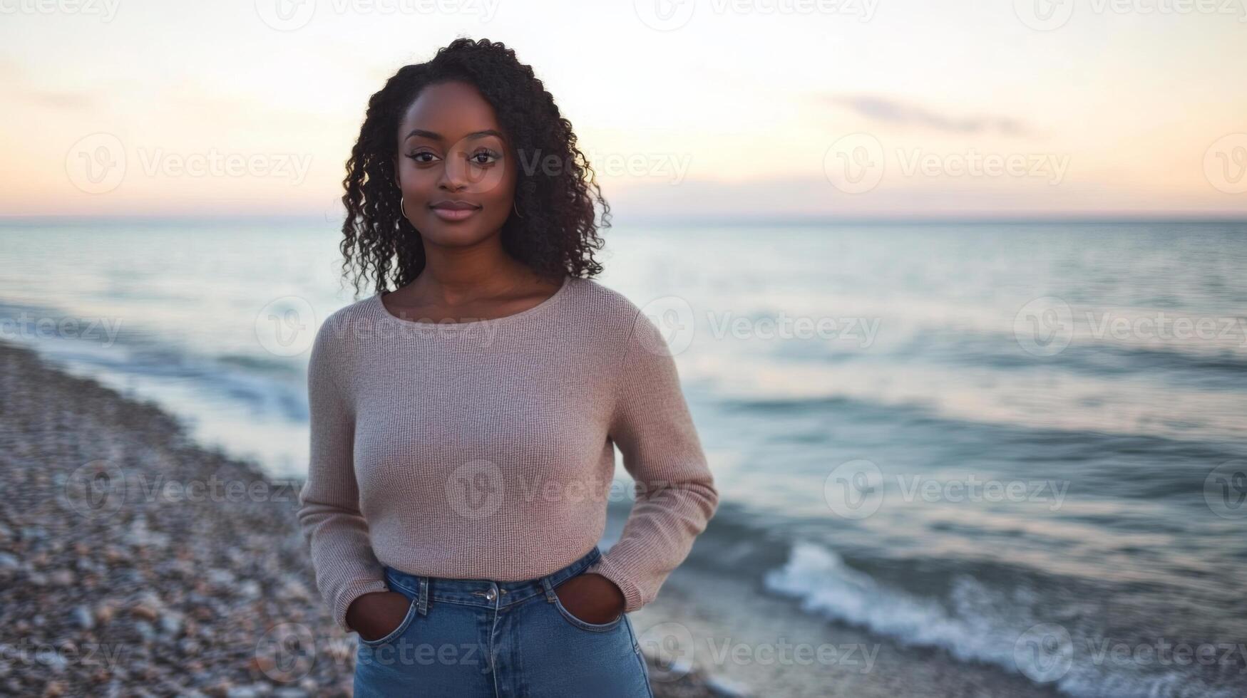 A young woman with natural curls stands confidently on a pebble beach during sunset. The sun casts a warm glow over the water, creating a serene atmosphere, perfect for a peaceful moment. photo