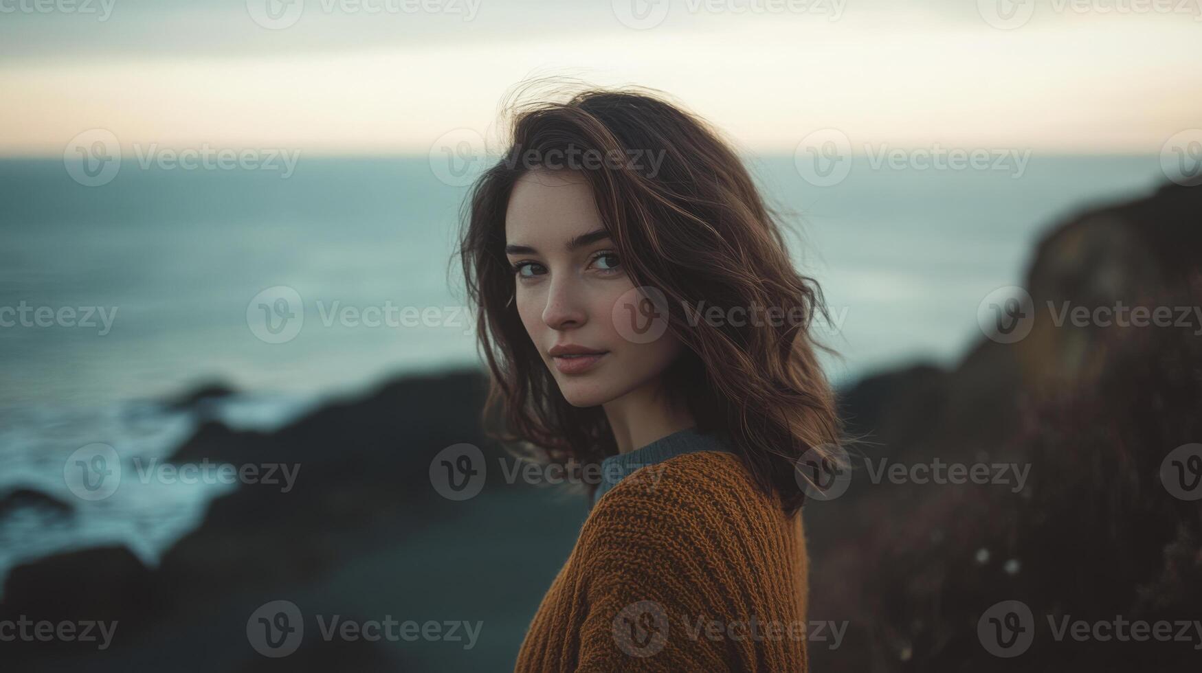 A young woman with wavy brown hair looks towards the ocean, dressed in a cozy sweater. The sun sets behind her, casting a warm glow on the coastline and waves crashing against the rocks. photo