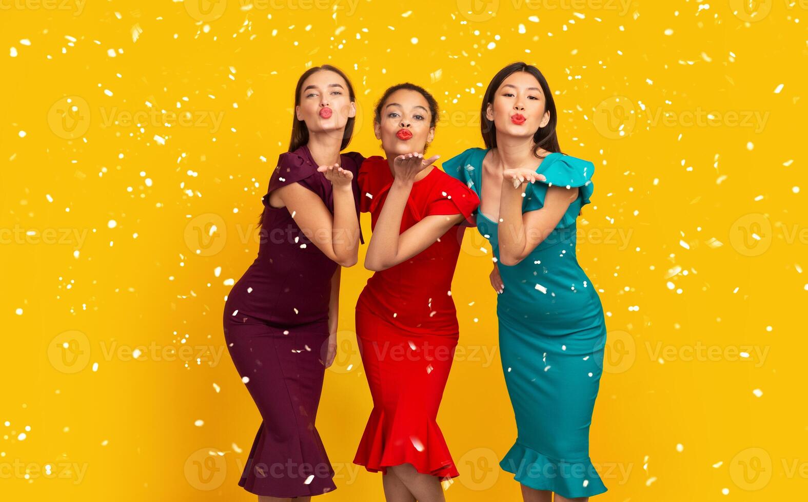 Three Diverse Girls In Colorful Dresses Sending Air Kiss To Camera Standing Over Yellow Background. Studio Shot photo