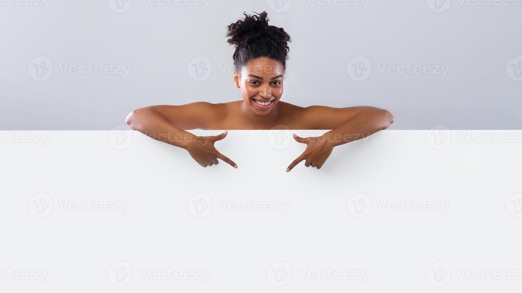 Bust of happy beautiful black nude girl pointing at empty space, grey background, spa, beauty salon advertisement photo