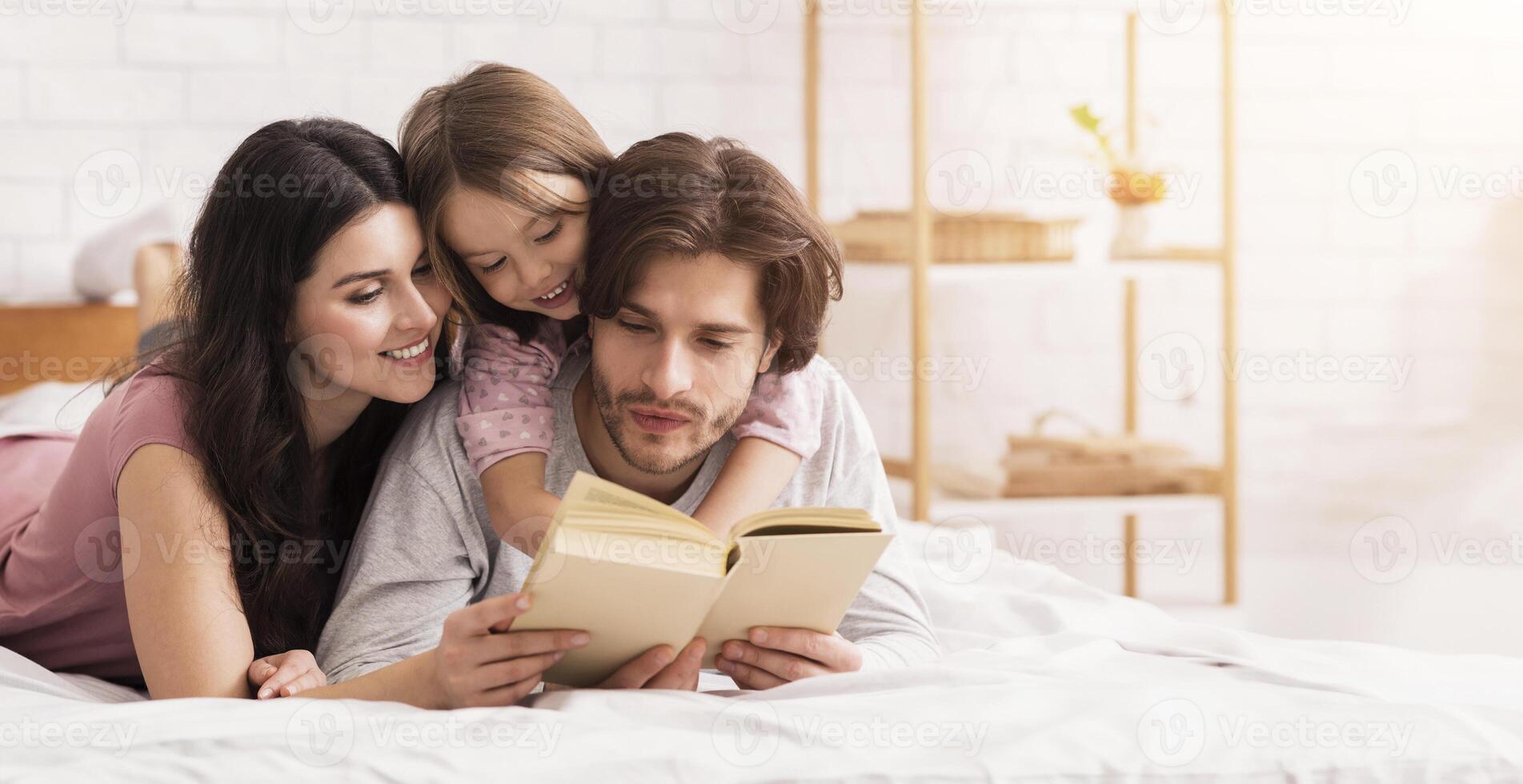 Loving Parents Reading Fairy Tale Book To Their Little Daughter, Lying On Bed Together And Embracing photo