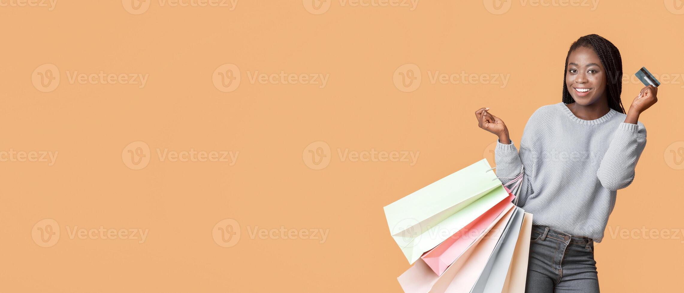 A young black woman stands joyfully, holding multiple shopping bags in pastel colors while displaying a credit card, copy space photo