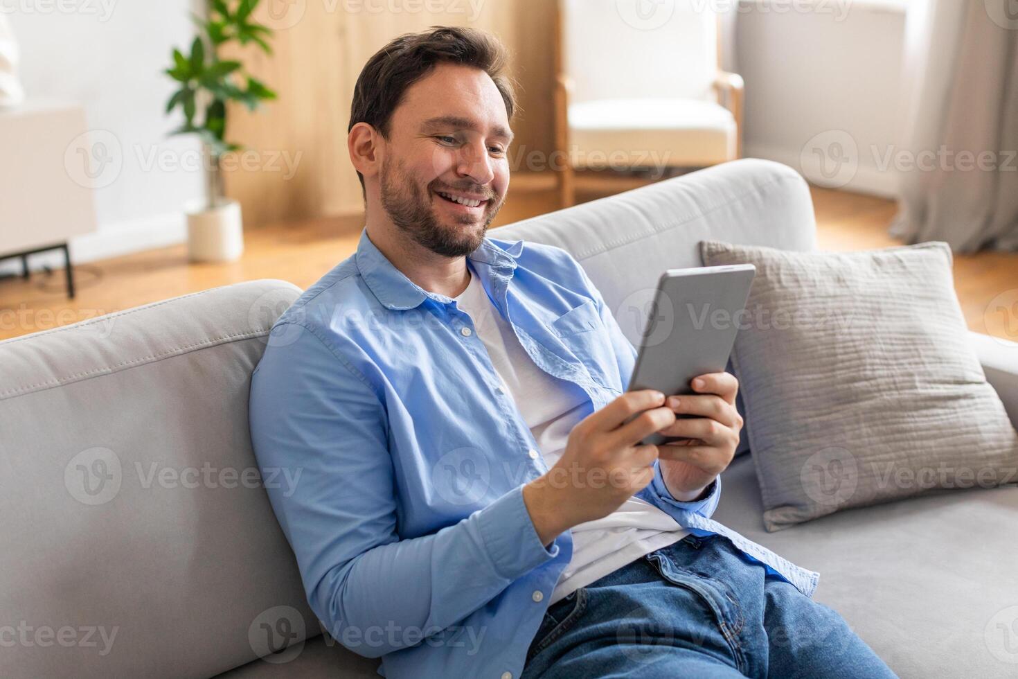 A man is seated on a couch, engrossed in using a tablet computer. He is focused on the screen, tapping and scrolling through content. The room is well-lit photo