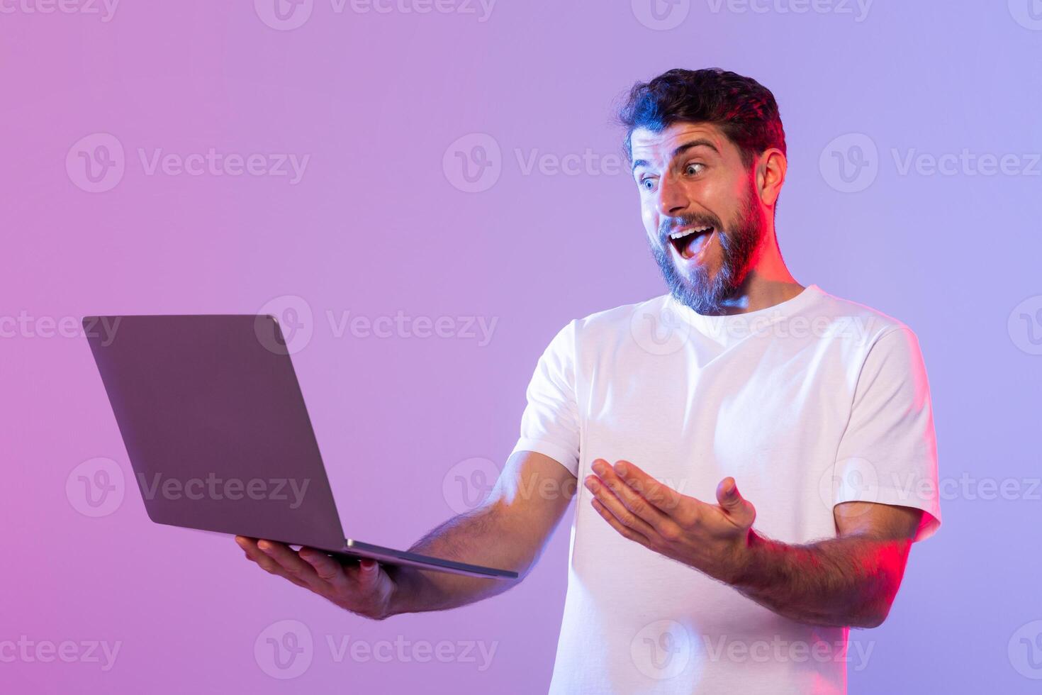 A young man with a beard and wearing a white t-shirt is holding a laptop with an excited and surprised expression on his face. The background features a gradient of purple and light blue photo