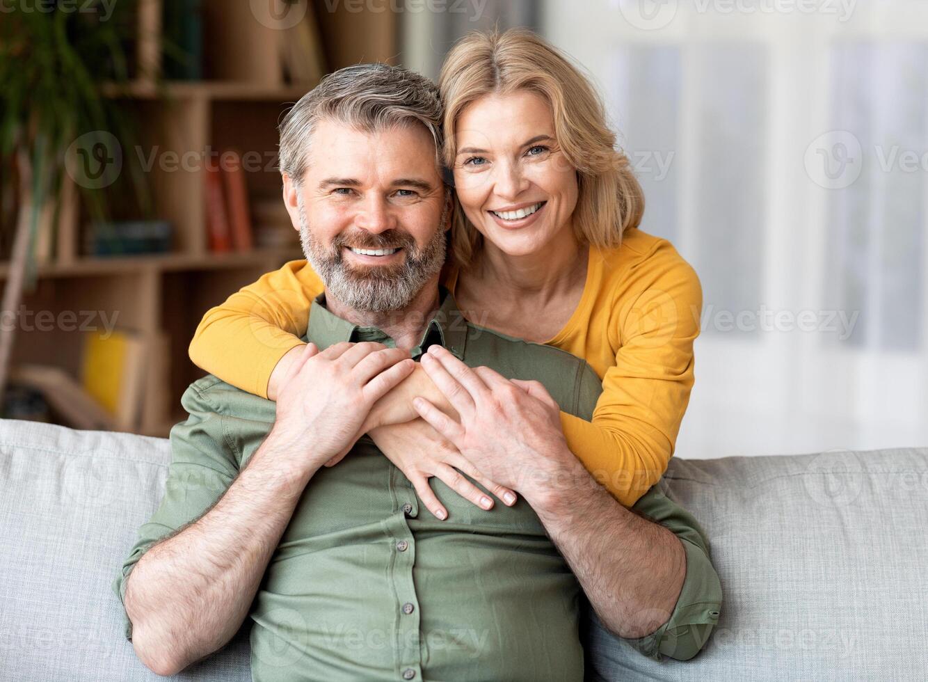 Portrait of beautiful smiling middle aged couple embracing at home, cheerful mature spouses relaxing together in living room while posing at camera, loving wife hugging her husband from behind photo