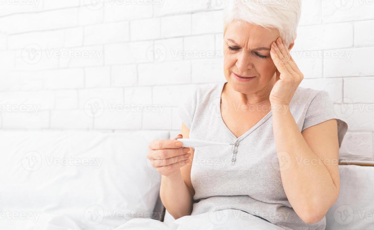 A senior woman with a concerned expression is sitting in her bed, holding a thermometer, appearing to check for a fever while touching her forehead as if experiencing a headache or flu symptoms. photo