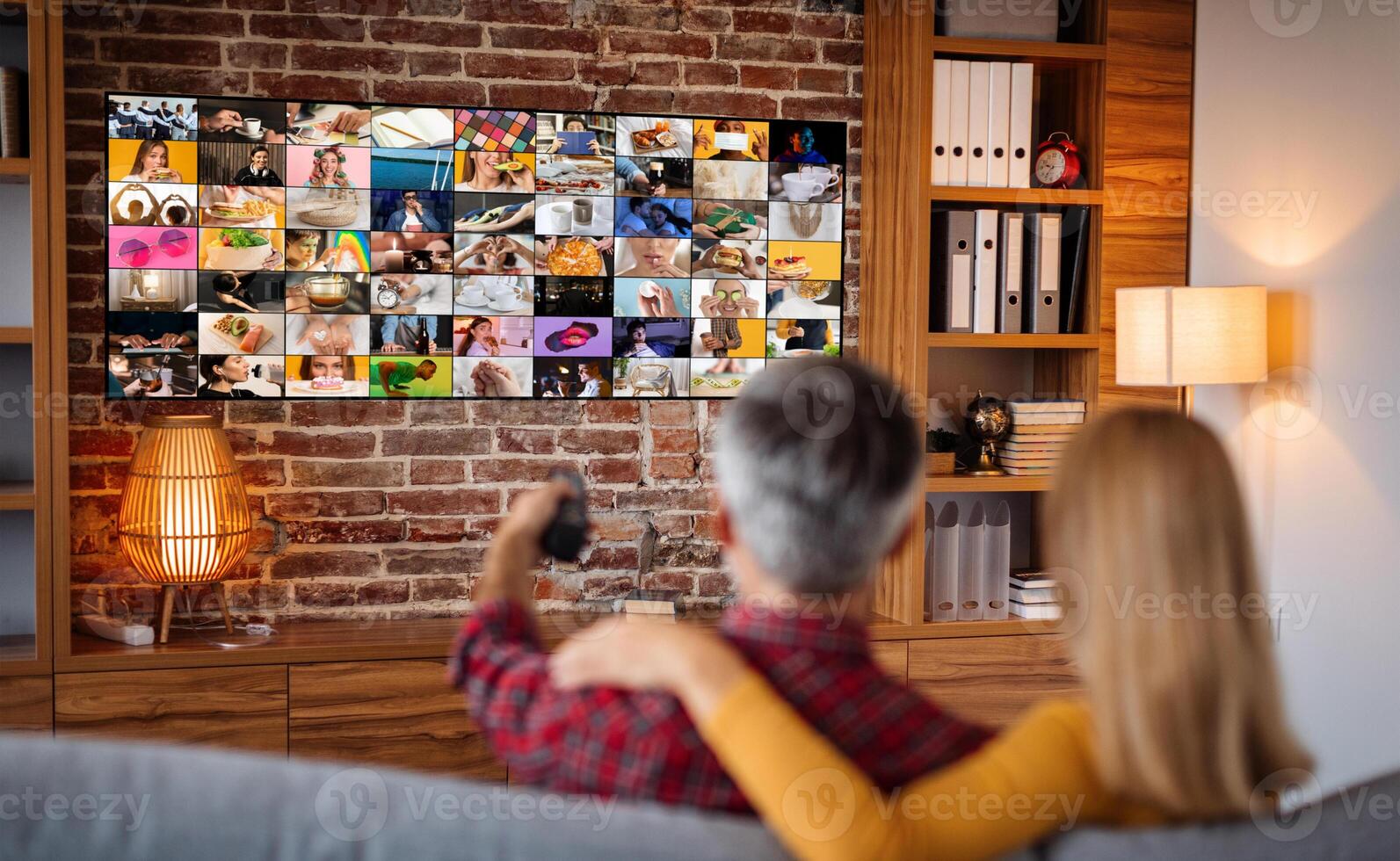 A couple is relaxing on a couch in their living room, watching a television screen filled with a collage of various images. The television is mounted on a brick wall photo