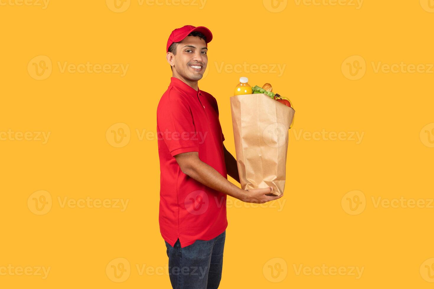 Delivery man standing over yellow studio background, holding a paper bag filled with various food items. He is looking directly at the camera photo