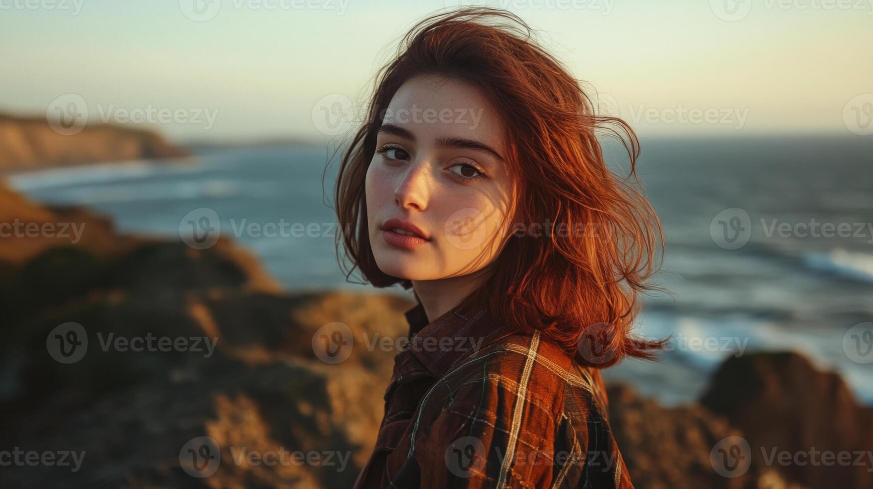 A young woman stands on a rocky cliff, looking out over the ocean's waves during golden hour. Her hair is gently tousled by the breeze as she enjoys the serene coastal landscape. photo