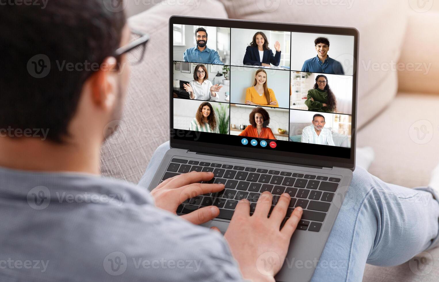 A young man wearing glasses sits on a sofa and takes part in a virtual team meeting via a laptop with eight colleagues appearing on the screen, during the afternoon. photo
