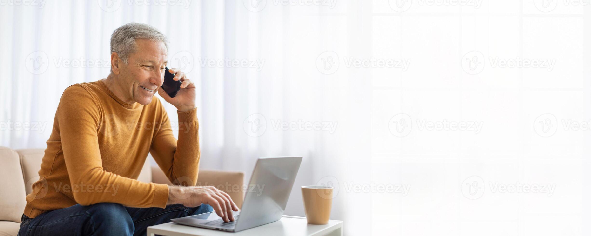 A mature man in a yellow sweater sits on a couch in a bright room while working on a laptop. He is talking on a cell phone and looks at the screen, copy space photo