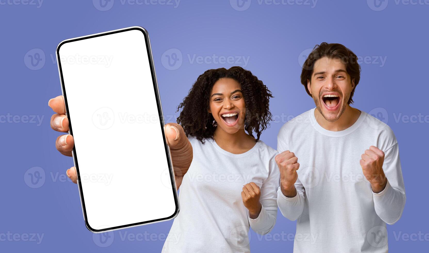 Multiracial couple celebrating their success while looking at a smartphone with a blank screen. The woman is holding the phone in front of her. The man smiling and raising his fists in the air photo